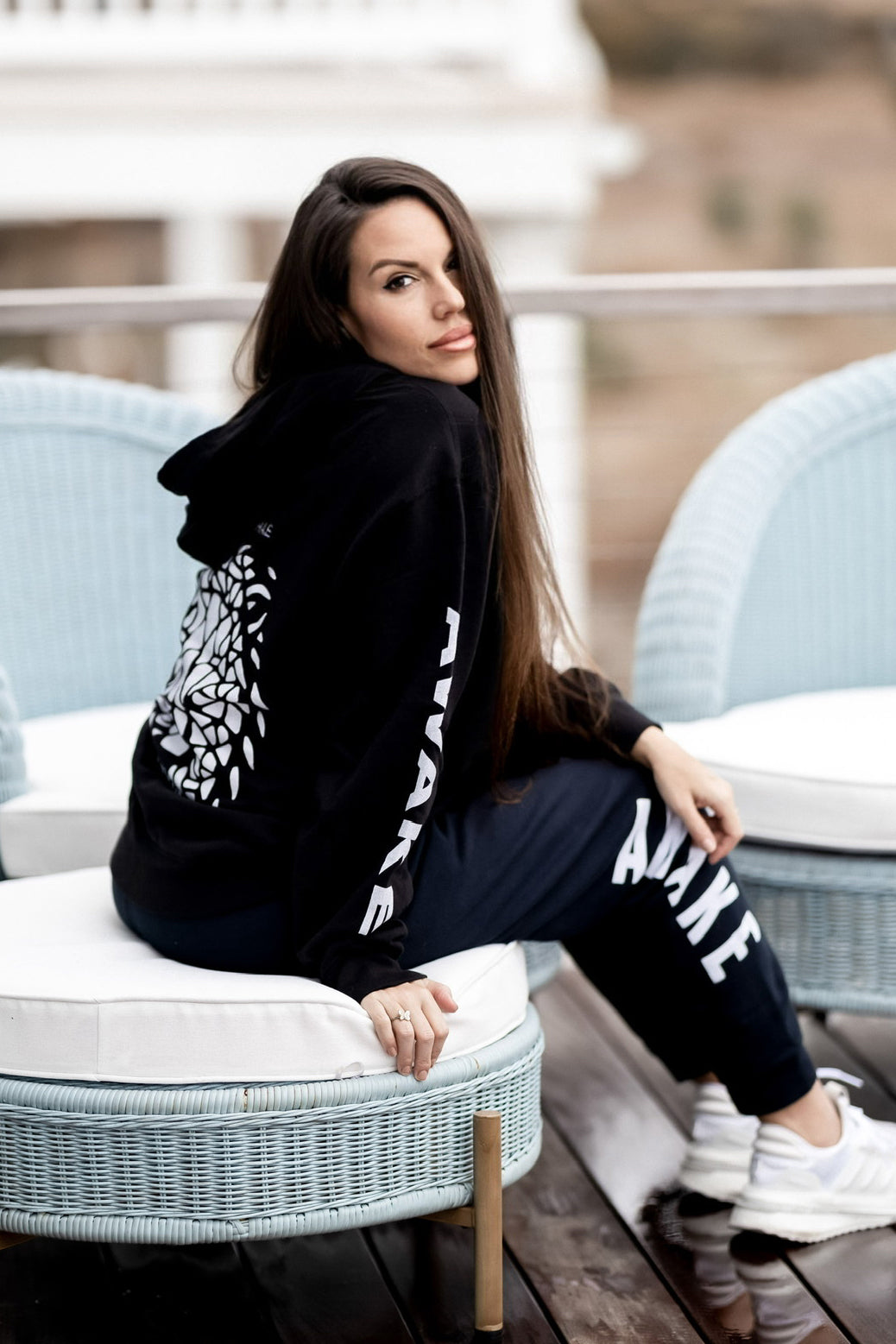 Beckah Shae wearing a black Awake Lion hoodie and Awake pants, sitting on a wicker chair outdoors.