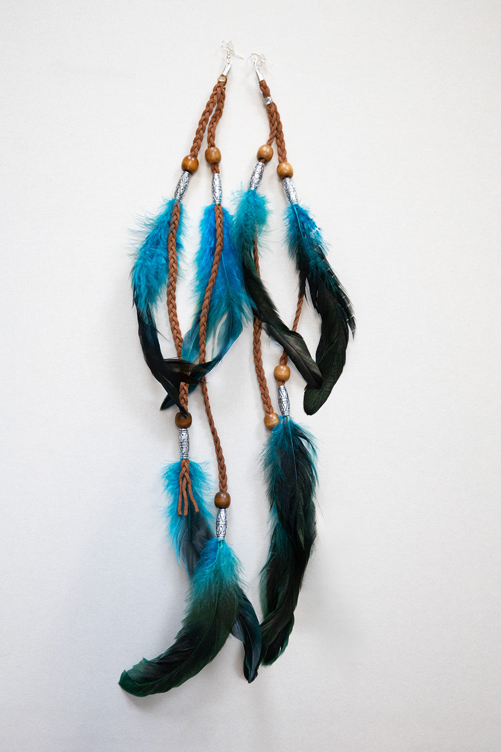 Covered Feather Earrings in teal and black feathers with braided brown leather, wooden beads, and silver accents, displayed on a white background.