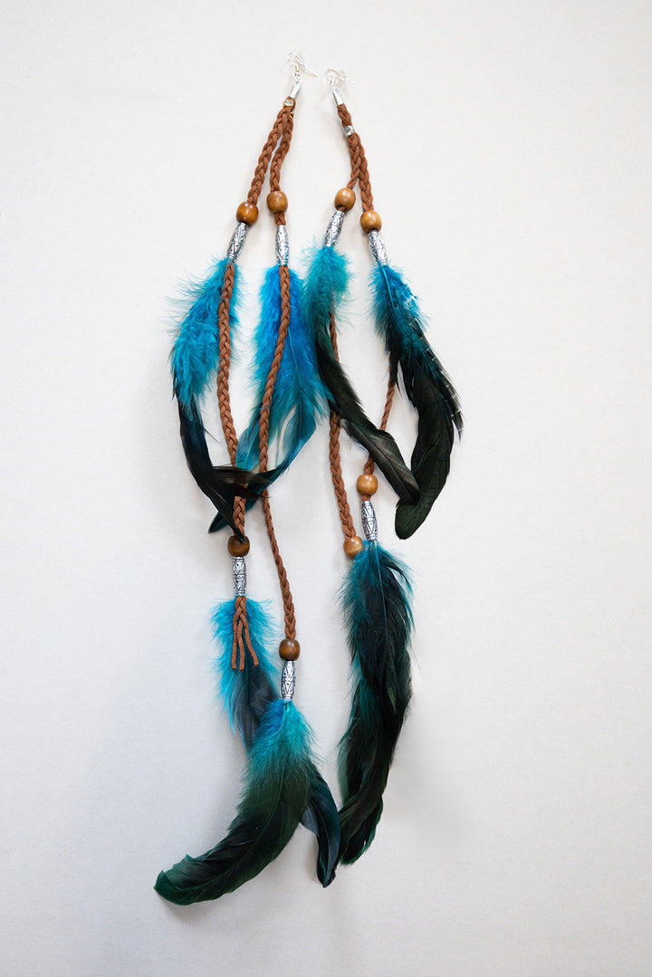 Covered Feather Earrings in teal and black feathers with braided brown leather, wooden beads, and silver accents, displayed on a white background.