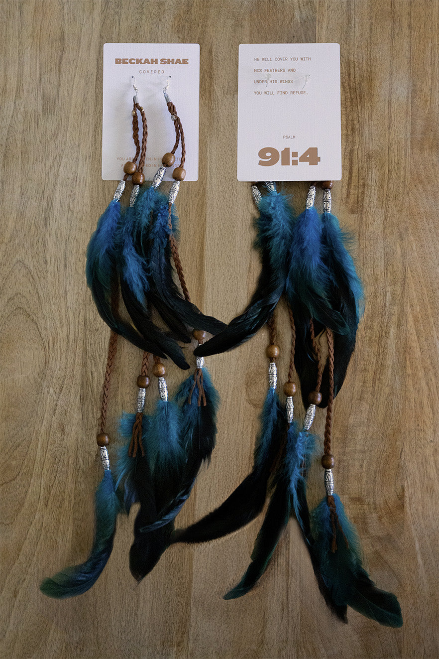 Beckah Shae Covered feather earrings with teal and black feathers, braided leather, wooden beads, and silver accents displayed on front and back cards with Psalm 91:4 scripture.