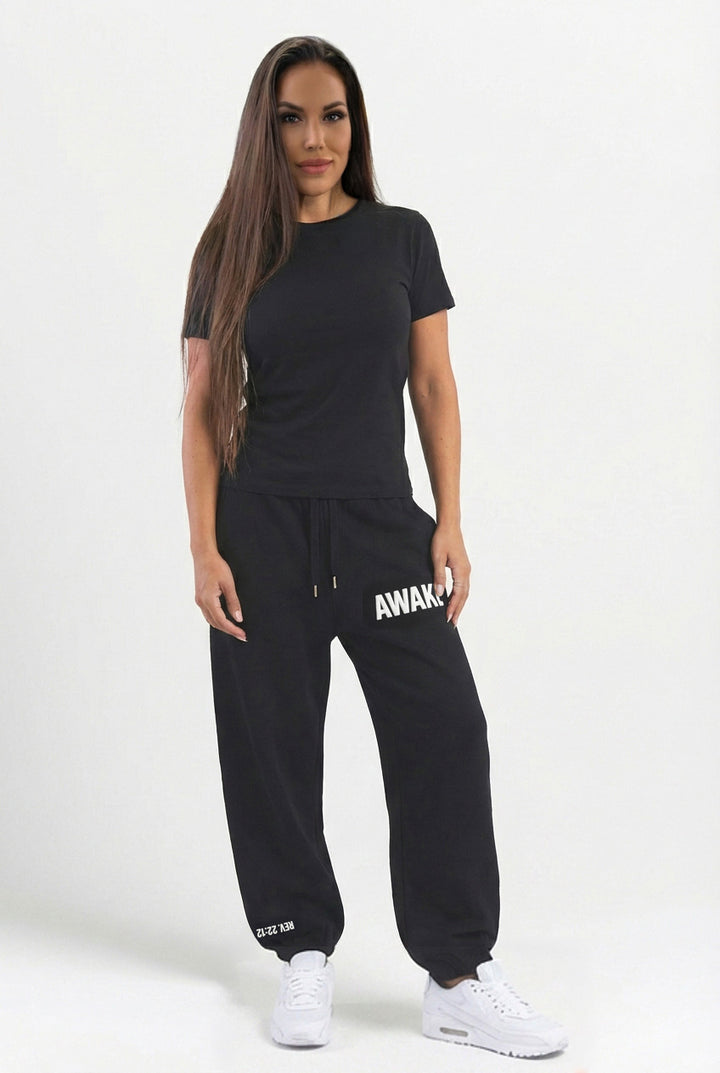 Woman wearing a black t-shirt and black sweatpants with 'AWAKE' branding on a white background with the scripture Revelation 22:12 on the bottom ankle.